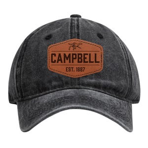 May include: A black, distressed baseball cap with a curved brim. The cap features a brown leather patch with the name "CAMPBELL" and "EST. 1887" in a serif font. A graphic of a running animal is above the name.