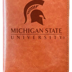 May include: A brown leather-bound journal with a bookmark. The front cover features a brown Spartan helmet logo and the text "MICHIGAN STATE UNIVERSITY".