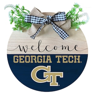 May include: A round wooden door hanger with a welcome sign and the Georgia Tech logo. The logo is gold and blue on a navy blue background. The top of the hanger is decorated with a burlap bow and white and black checkered ribbon.