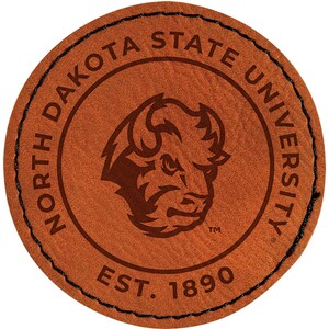May include: A round, brown leather patch with the words "NORTH DAKOTA STATE UNIVERSITY" encircling a bison head logo. The text "EST. 1890" is at the bottom. The edge is stitched with black thread.