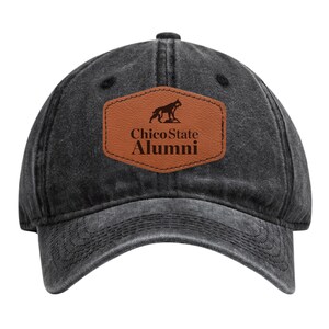 May include: A black baseball cap with a curved brim. The cap features a brown leather patch with the text "Chico State Alumni" and a silhouette of a dog. The cap is made of a faded, distressed material.