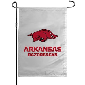 May include: A white garden flag featuring the Arkansas Razorbacks logo. The logo includes a red Razorback hog and the words "ARKANSAS RAZORBACKS" in red lettering. The flag is hanging from a black metal pole.