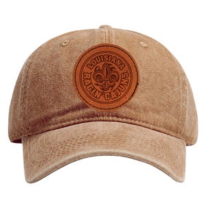 May include: A tan baseball cap with a curved brim. A circular brown leather patch on the front displays the words "Louisiana Ragin' Cajuns" around a fleur-de-lis symbol. The cap is made of a soft, worn material.