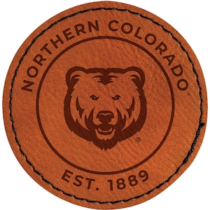 May include: A round, brown leather patch with the words "NORTHERN COLORADO" arched around the top. A bear's head is in the center. The text "EST. 1889" is at the bottom. The edge is stitched with black thread.