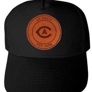 May include: Black trucker hat featuring a brown leather patch with the text "UC DAVIS" and "EST. 1908". The patch also includes a stylized letter "A" inside a curved shape. The hat has a mesh back and a curved brim.