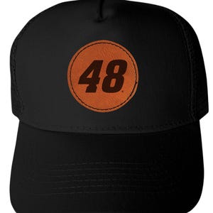 May include: Black trucker hat with a curved brim and mesh back. The front features a circular patch with the number "48" in brown against an orange background. The hat has a snap closure.