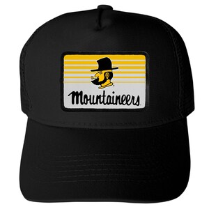 May include: A black trucker hat featuring a rectangular patch on the front. The patch displays horizontal yellow and white stripes with a graphic of a bearded person wearing a hat and smoking a pipe, above the text Mountaineers in black script.
