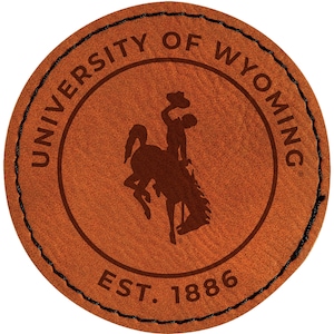 May include: A round, brown leather patch with the words "UNIVERSITY OF WYOMING" in a circular pattern. The center features a silhouette of a cowboy riding a bucking bronco. The text "EST. 1886" is at the bottom. The edge is stitched with black thread.