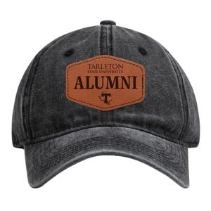 May include: A black, distressed baseball cap with a curved brim. A brown leather patch on the front displays the text "TARLETON STATE UNIVERSITY ALUMNI" in a serif font, with a "T" logo below. The cap is made of a worn-looking fabric.