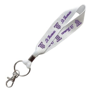 May include: A white fabric lanyard featuring a repeating purple St. Thomas logo and text pattern. The lanyard includes a silver-toned metal crimp, a circular split ring, and a spring-loaded lobster claw clasp for attaching keys or identification badges.