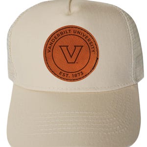 May include: Cream-colored trucker hat with a curved brim and mesh back. The front features a circular brown leather patch with the words "Vanderbilt University" and a "V" logo. The hat is a classic style, suitable for casual wear.