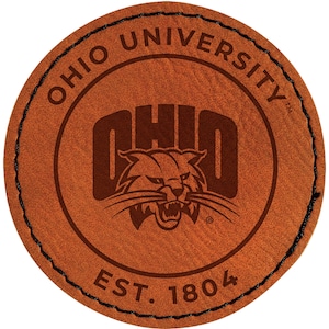 May include: A round, brown leather patch with the words "OHIO UNIVERSITY" arched around the top. The center features the word "OHIO" and a wildcat logo. The text "EST. 1804" is at the bottom. The edge is stitched with black thread.