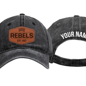 May include: Two dark gray baseball caps. One cap features a brown leather patch with the word "REBELS" and "EST. 1997". The other cap has the text "YOUR NAME" in white. Both caps have an adjustable strap.