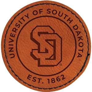 May include: A round, brown leather patch with the words "UNIVERSITY OF SOUTH DAKOTA" in a circular pattern. The center features the letters "SD" in a stylized design. The text "EST. 1862" is at the bottom. The patch has a stitched black border.