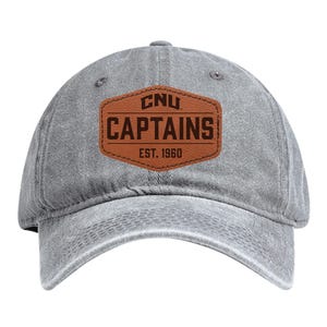 May include: A gray baseball cap with a curved brim. The front features a brown leather patch with the text "CNU CAPTAINS EST. 1960" in white and brown lettering. The cap is made of a soft, worn material.