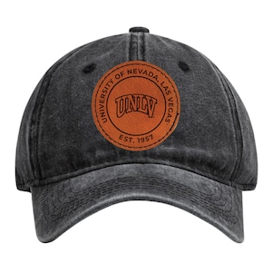 May include: A black, distressed baseball cap featuring a circular leather patch. The patch displays the text "UNIVERSITY OF NEVADA, LAS VEGAS" around the edge, with "UNLV" in the center and "EST. 1957" below. The cap has a curved brim.