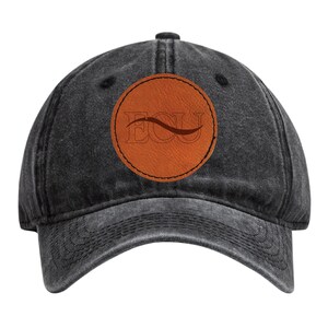 May include: A black, washed-out baseball cap with a curved brim. The cap features a circular, brown leather patch with the letters "ECU" and a curved line design. The hat is made of a soft, worn material.