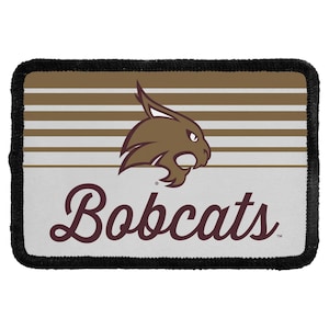 May include: A rectangular patch with a black stitched border. The top half features horizontal brown and white stripes with a brown bobcat head graphic. The bottom half is light gray with the word Bobcats written in a dark brown script font.