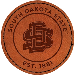 May include: A round, brown leather patch with the words "SOUTH DAKOTA STATE" in a circular pattern. The center features a stylized "SDS" logo in a darker brown, with "EST. 1881" below. The edge is stitched with black thread.