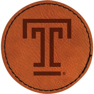 May include: A round, brown leather patch with a stitched black border. The patch features a dark brown letter "T" in the center. The design includes a registered trademark symbol.