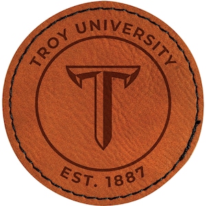 May include: A round, brown leather patch with the words "TROY UNIVERSITY" in a circular pattern around the top. The center features a stylized "T" within a shield shape. The text "EST. 1887" is at the bottom. The edge is stitched with black thread.