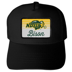 May include: A black baseball cap featuring a rectangular patch on the front. The patch displays the green and yellow NDSU logo with a bison graphic above the word Bison in green script text, set against a background of horizontal yellow stripes.