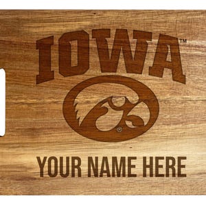 May include: A rectangular wooden cutting board with a natural grain finish and a handle slot on the left side. The board features the engraved text IOWA above a stylized hawk logo and the placeholder text YOUR NAME HERE at the bottom.