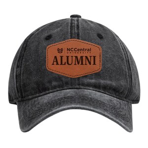 May include: A black, distressed-look baseball cap with a curved brim. The cap features a brown leather patch with the text "NC Central University ALUMNI" in white lettering. The cap is made of a durable fabric and has an adjustable strap.
