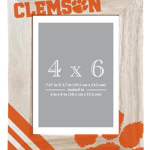 May include: Wooden photo frame with the word "CLEMSON" in orange, along with a paw print design. The frame is designed for a 4 x 6 inch photo, with the overall frame dimensions of 7.67 x 5.7 inches. The frame has orange stripes.