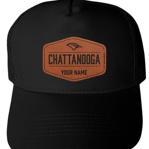 May include: A black trucker hat featuring a brown, hexagonal leather-like patch on the front. The patch displays a bird icon, the text CHATTANOOGA, and the text YOUR NAME in black lettering. The hat has a solid front panel and mesh back panels.