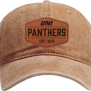 May include: A tan baseball cap with a curved brim. The cap features a brown leather patch with the text "UNI PANTHERS EST. 1876" in black. The cap has a distressed look.