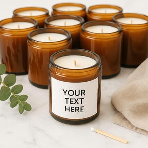May include: An assortment of amber glass jar candles with white wax and wooden wicks. One candle features a white label with the text "YOUR TEXT HERE". A match and some greenery are also visible.