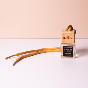 May include: A clear glass diffuser bottle with a wooden cap and a mustard-colored cord. The label reads "Purpose White Tea Diffuser" and "Shine & Love Candles". The diffuser is on a white surface with a light pink background.