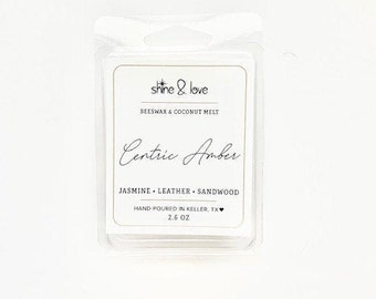 Op de afbeelding: Een witte was smelt met de tekst "shine & love" en "Beeswax & Coconut Melt" in zwarte tekst. De tekst "Centric Amber" is in zwarte cursieve tekst. De tekst "Jasmine - Leather - Sandwood" is in zwarte tekst. De tekst "Hand-poured in Keller, TX" en "2.6 oz" is in zwarte tekst.