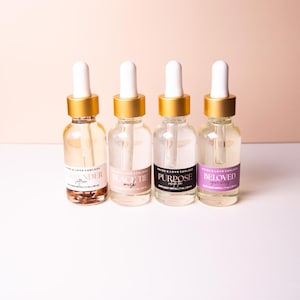 Könnte beinhalten: Vier Glas-Tropfflaschen mit goldenen Deckeln und weißen Tropfern. Die Flaschen sind mit den Düften Lavender, Black Tie, Purpose und Beloved beschriftet. Die Flaschen sind mit klarer Flüssigkeit gefüllt.