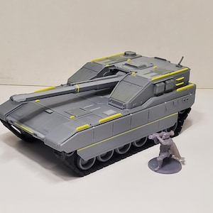 May include: A gray plastic model tank featuring a long main cannon, angular hull design, and bright yellow accent strips along the top and sides. Beside the tank stands a small, unpainted gray miniature figure holding a rocket launcher weapon.