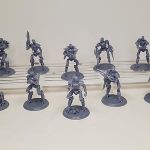 Op de afbeelding: Een verzameling van tien grijze robotfiguren, elk op een cirkelvormige basis. De robots staan in verschillende poses, sommige met wapens. De figuren zijn gerangschikt op een wit lattenoppervlak.