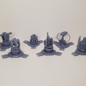 Könnte beinhalten: Eine Sammlung grauer Miniaturtürme, jede mit einem einzigartigen Design, darunter Kanonen und andere Waffensysteme. Die Türme sind auf kreisförmigen Sockeln mit rechteckigen Erweiterungen montiert.