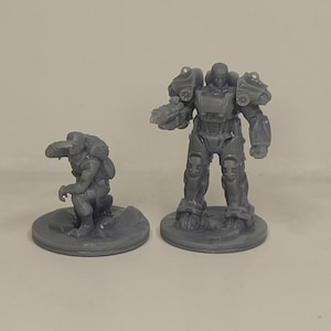 May include: Two grey miniature figurines. One is a crouching creature with a backpack, the other is a standing figure in futuristic armour. Both are on circular bases, suitable for tabletop gaming or display.