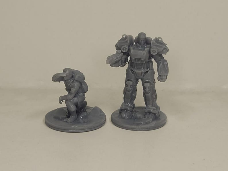 May include: Two grey miniature figurines. One is a crouching creature with a backpack, the other is a standing figure in futuristic armour. Both are on circular bases, suitable for tabletop gaming or display.