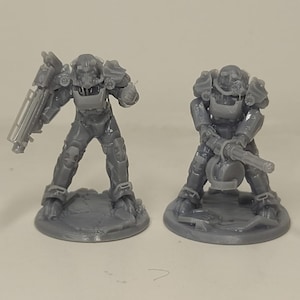 May include: Two grey miniature figurines of armoured characters, one holding a large weapon and the other with a similar weapon. The figures are standing on circular bases, with detailed armour and helmets.