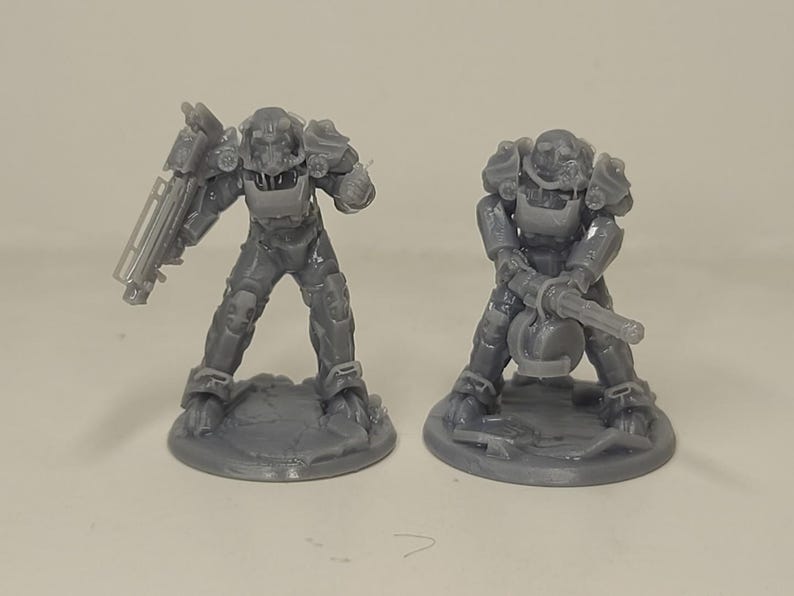May include: Two grey miniature figurines of armoured characters, one holding a large weapon and the other with a similar weapon. The figures are standing on circular bases, with detailed armour and helmets.