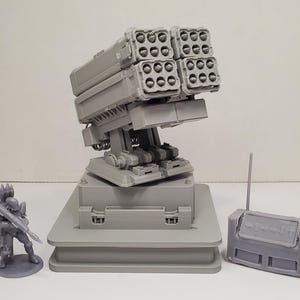 May include: A gray 3D printed miniature set featuring a large multi-tube rocket launcher system on a square base, a small soldier figurine holding a shoulder-mounted weapon, and a rectangular control console unit with a vertical antenna.