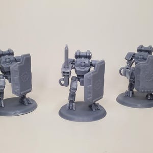 Puede incluir: Tres figuras de robots grises, cada una sobre una base circular. Los robots tienen diseños mecánicos detallados, uno sosteniendo un arma, otro una espada, y todos con escudos con un engranaje.