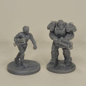 May include: Two grey miniature figurines. One is a soldier with a helmet and weapon, the other is a larger figure in armour, also holding a weapon. Both are standing on circular bases.