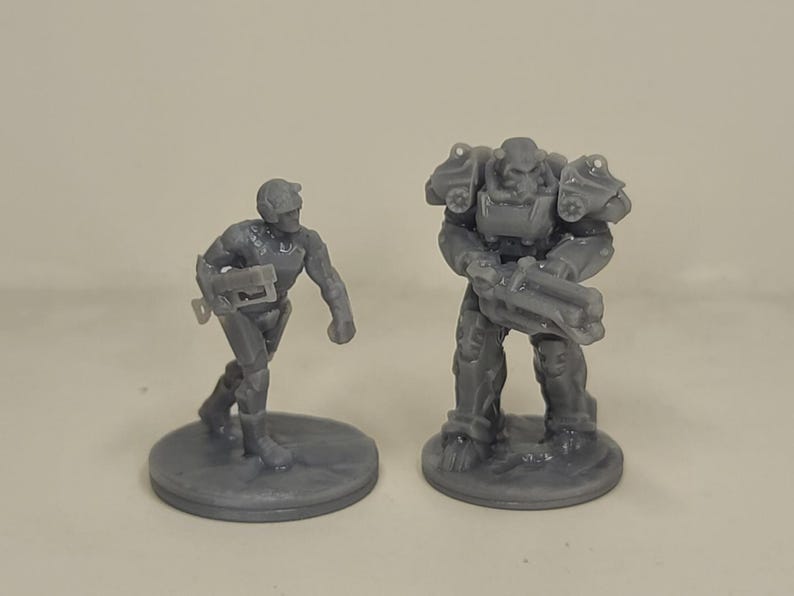 May include: Two grey miniature figurines. One is a soldier with a helmet and weapon, the other is a larger figure in armour, also holding a weapon. Both are standing on circular bases.