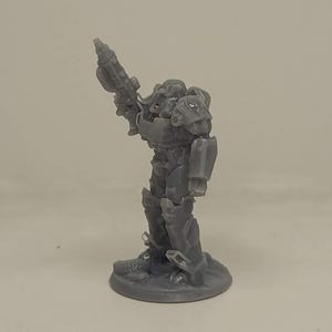 May include: A grey miniature figurine of a futuristic soldier in armour, holding a weapon. The figure is standing on a circular base. The armour has detailed panels and a helmet. The overall design suggests a science fiction or fantasy theme.