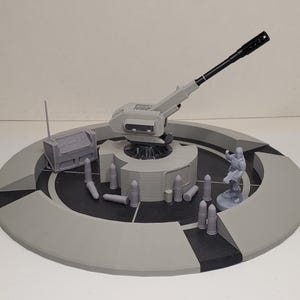 May include: A grey 3D printed circular platform featuring a central mounted artillery cannon with a long black barrel, a small rectangular communications box, several scattered shell casings, and a single standing soldier figurine.