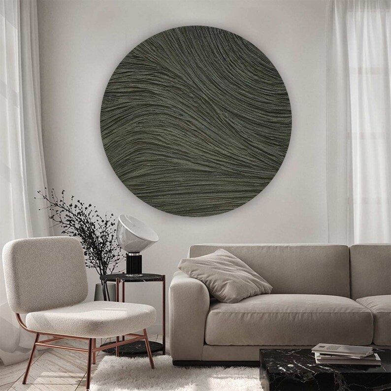 May include: A large, round, textured wall art piece in shades of gray and green hangs above a beige sofa. The artwork features a rippled, wave-like pattern. A modern chair with a copper frame and a small side table with a lamp are also visible.
