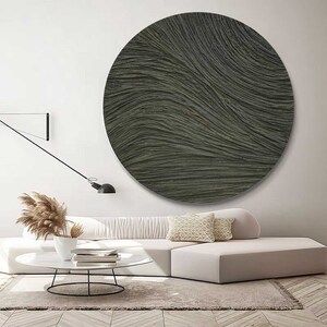 May include: A large, round, textured wall art piece in shades of gray and olive green hangs above a white sofa. The artwork features a rippled, wave-like pattern. The room is decorated in neutral tones with a modern aesthetic.